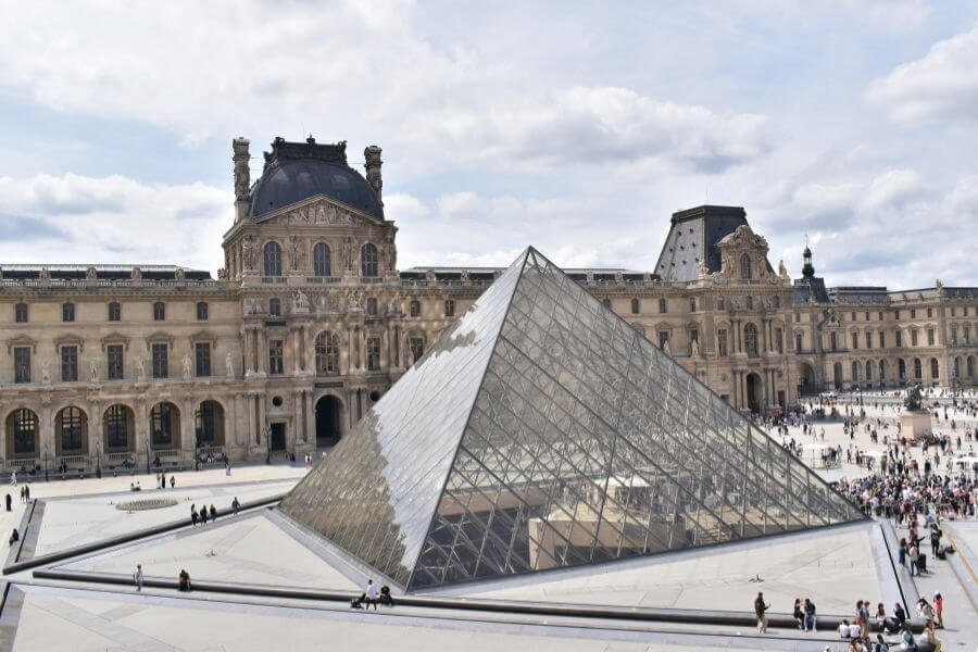 outside of the louvre with iconic glass pyramid, crowds sitting and wandering outside.
